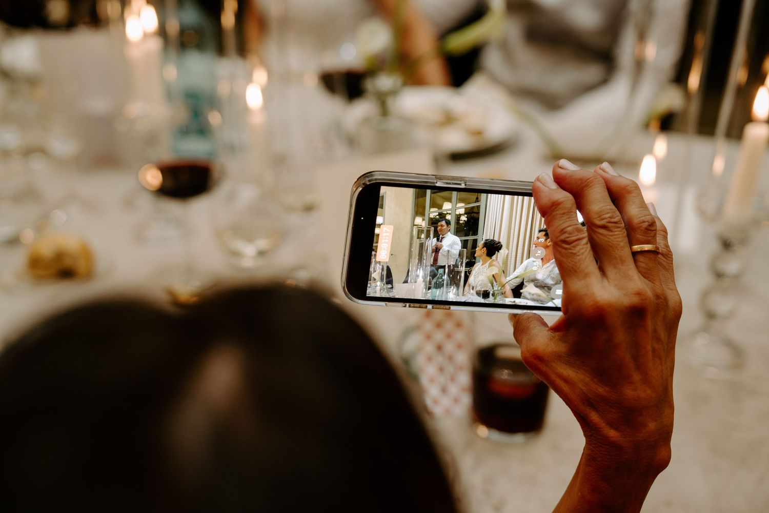 Wedding guest captures bride and groom on smartphone during candlelit reception dinner.