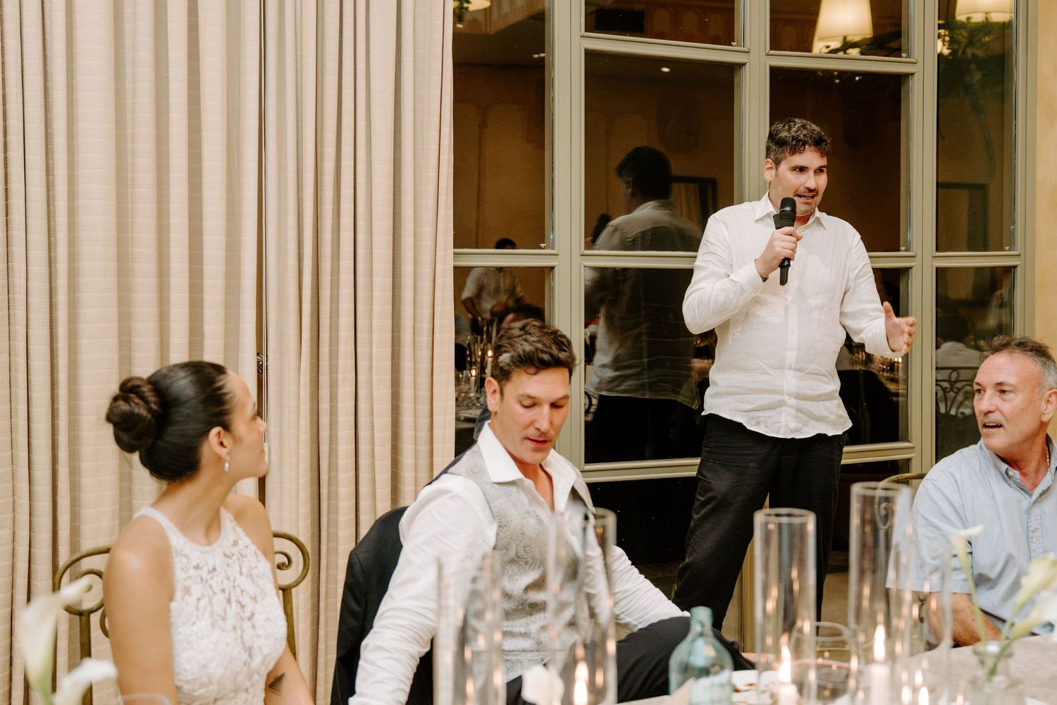 Best man delivers a heartfelt wedding speech with microphone while bride and groom listen at the table.