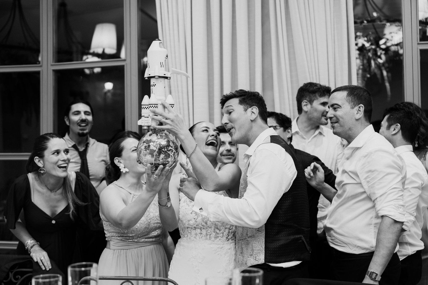 Joyful wedding guests surround bride and groom lifting their tiered wedding cake in a lively black and white photo.