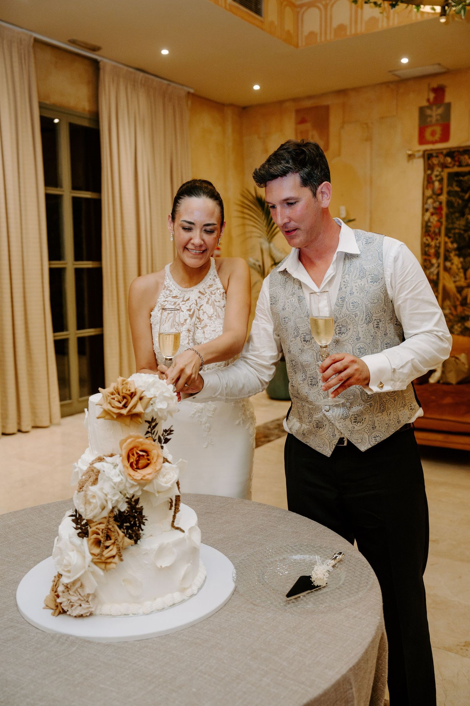 Bride and groom cutting their elegant white floral wedding cake while holding champagne glasses at their reception.