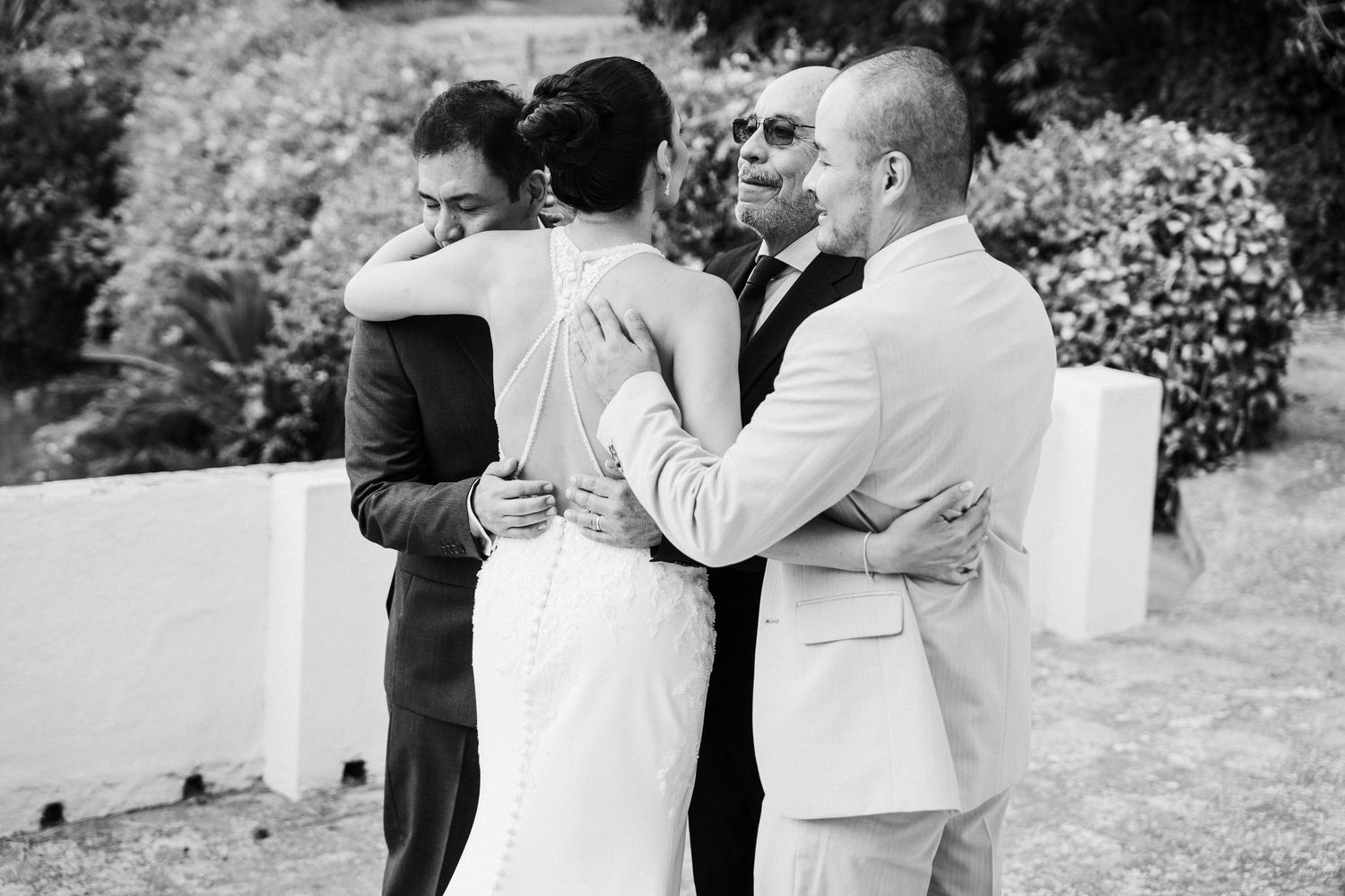Bride and groom share emotional embraces with family members outdoors in this black and white wedding photo.