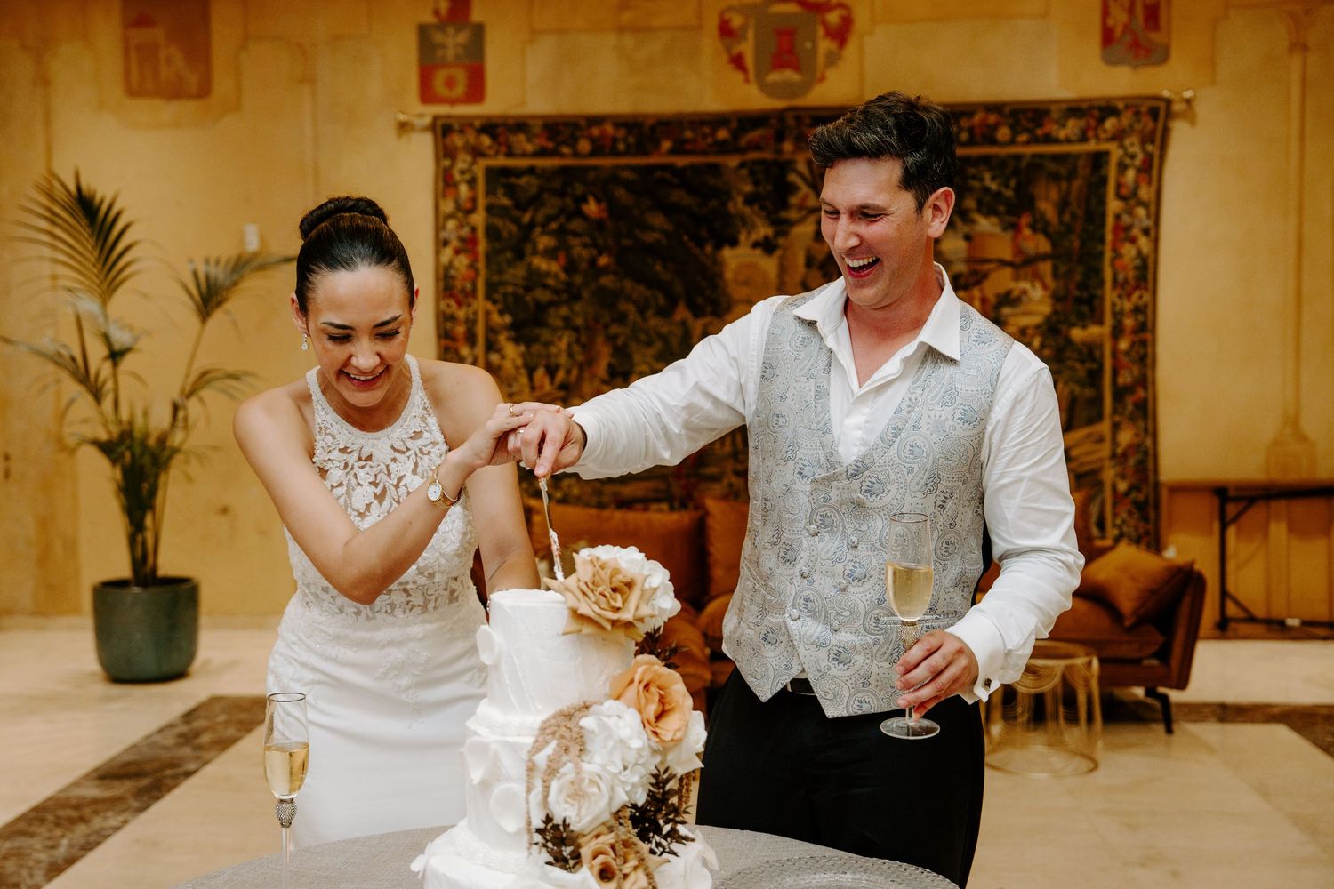 Bride and groom laughing while cutting elegant white wedding cake with floral decorations in ornate venue.