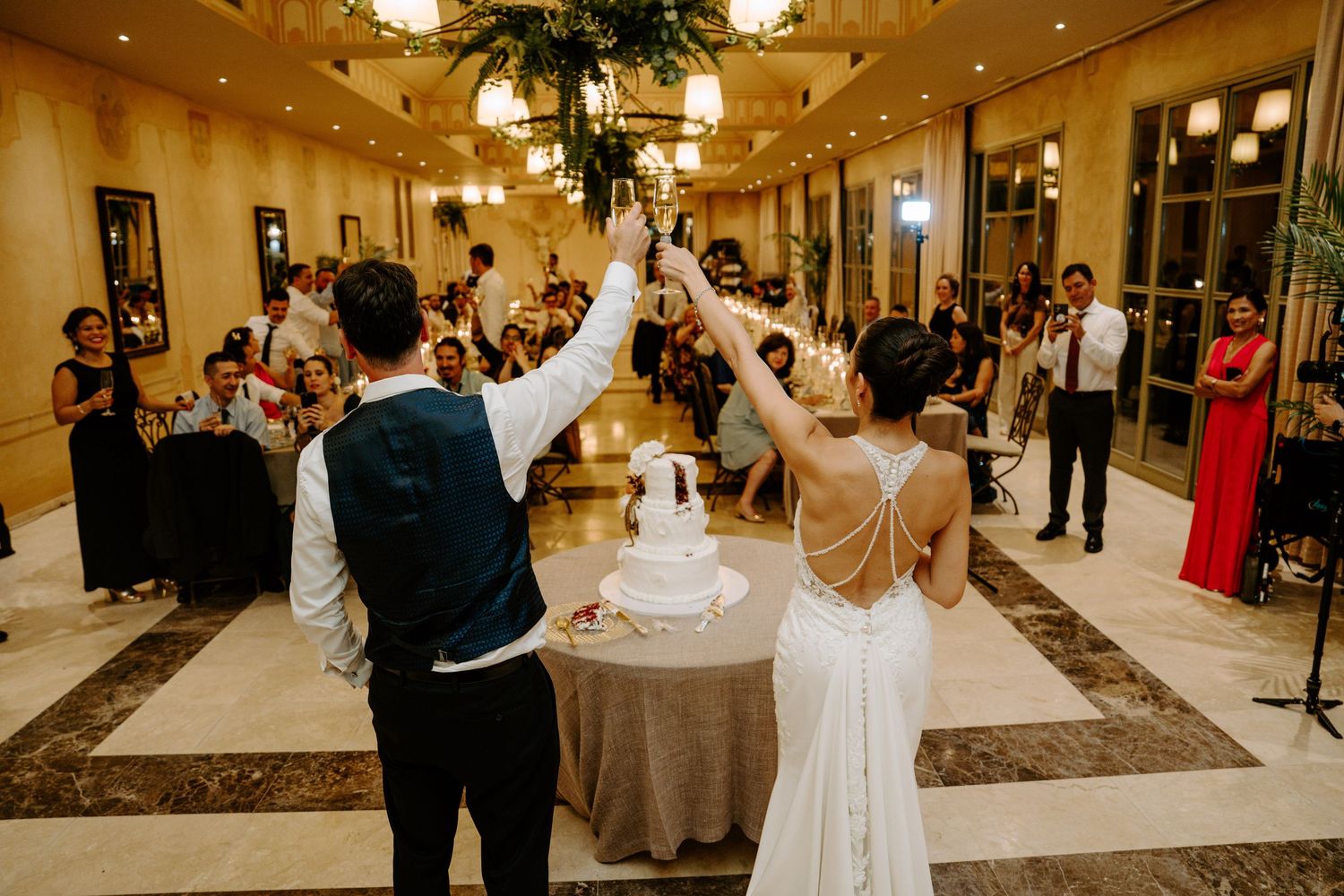Bride and groom raising glasses over wedding cake at elegant indoor reception with guests watching.