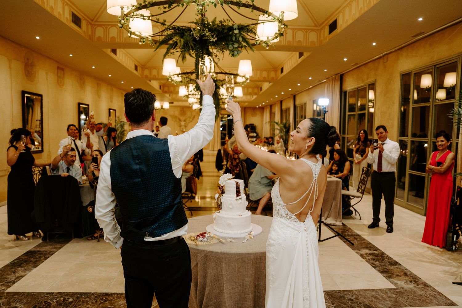 Bride and groom raising hands over wedding cake during reception while guests watch in elegant ballroom.