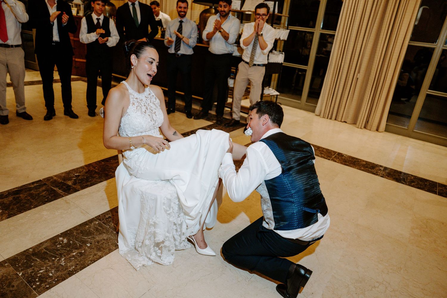 Groom kneeling before bride removing garter at wedding reception while guests celebrate in background.