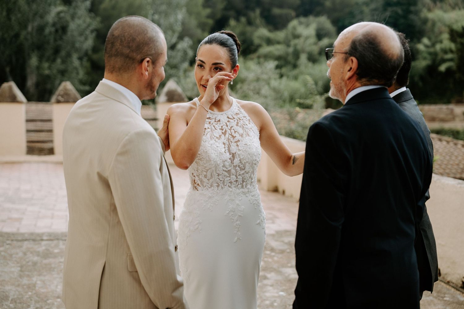 Emotional bride wiping tears while groom and officiant stand nearby during outdoor wedding ceremony.