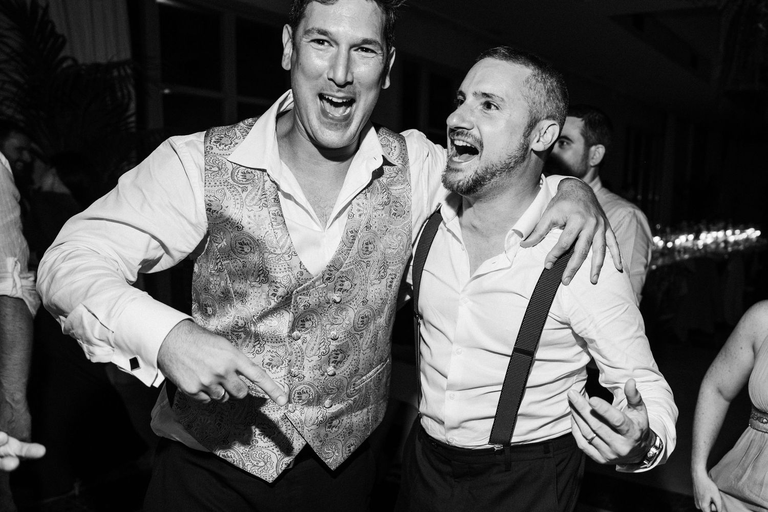 Two men laughing and dancing together in formal attire at a wedding reception, captured in black and white.