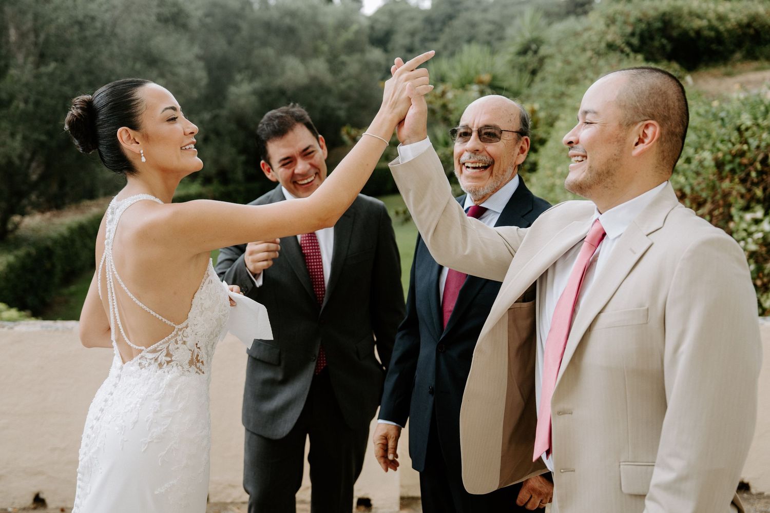 Bride and groom share a high-five with smiling groomsmen in pink ties at an outdoor wedding.