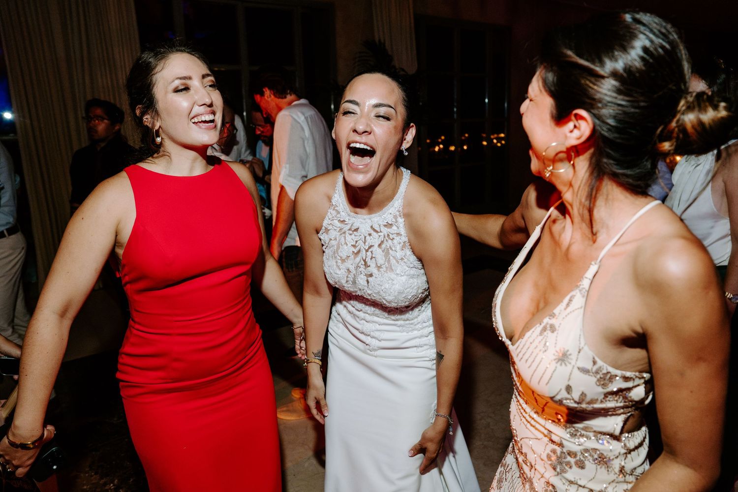 Three women laughing and celebrating together at a wedding reception, one wearing a red dress and another in a white bridal gown.