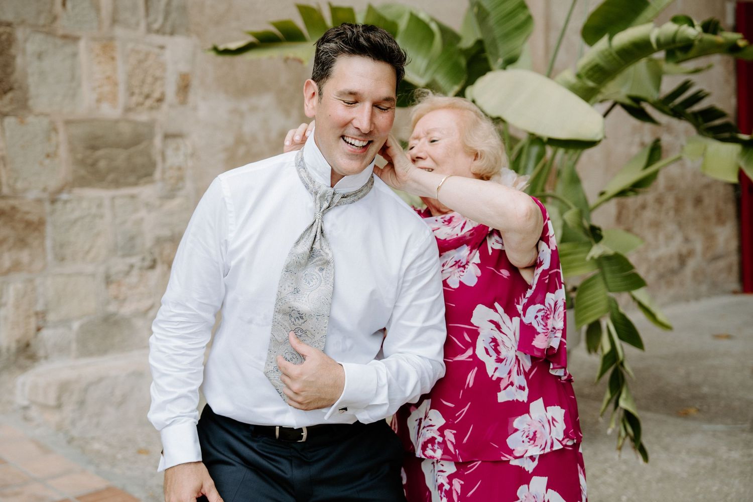 Joyful man in white shirt and tie laughs with older woman in pink floral dress at outdoor wedding.