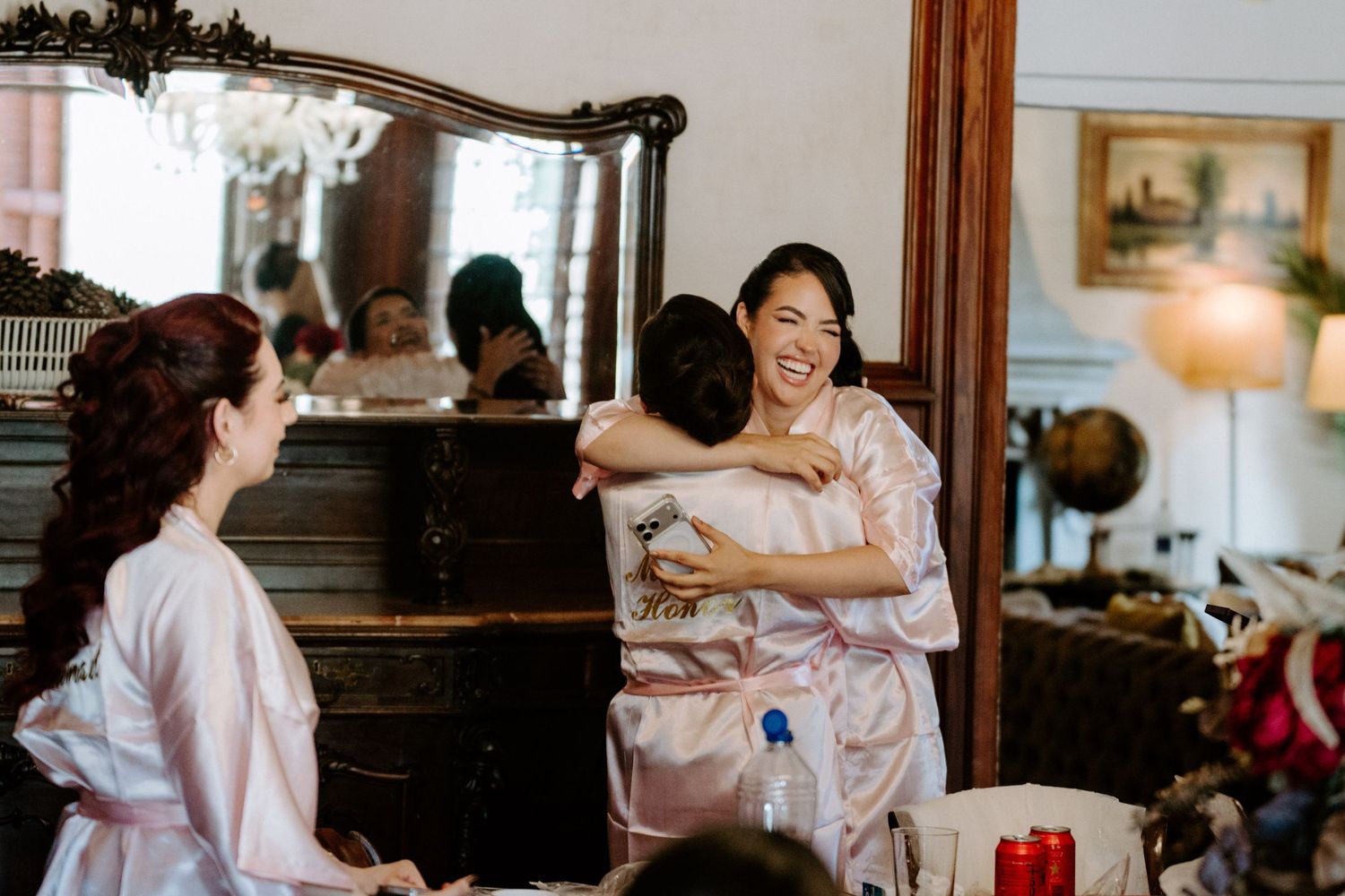 Bridesmaids in pink robes share a joyful hug during wedding preparations in an elegant vintage room.
