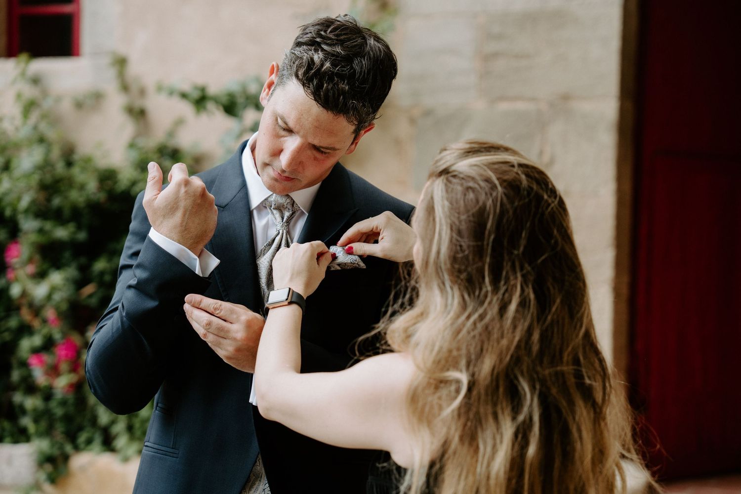 Groom in navy suit getting boutonniere pinned by a woman outdoors at a rustic wedding venue.