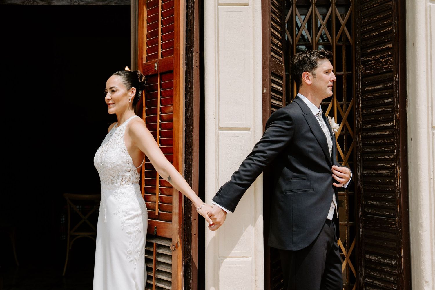 Bride and groom holding hands in doorway, bride in fitted lace gown and groom in dark suit.