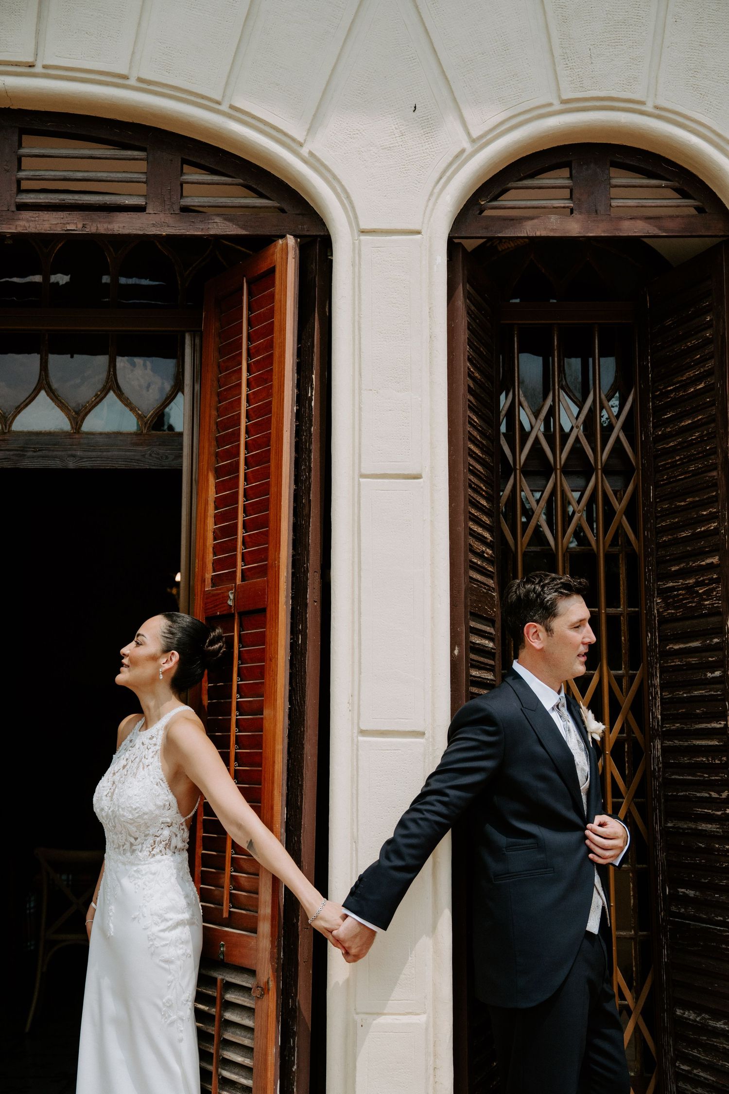 Bride and groom hold hands facing opposite directions by ornate wooden doors at their wedding.