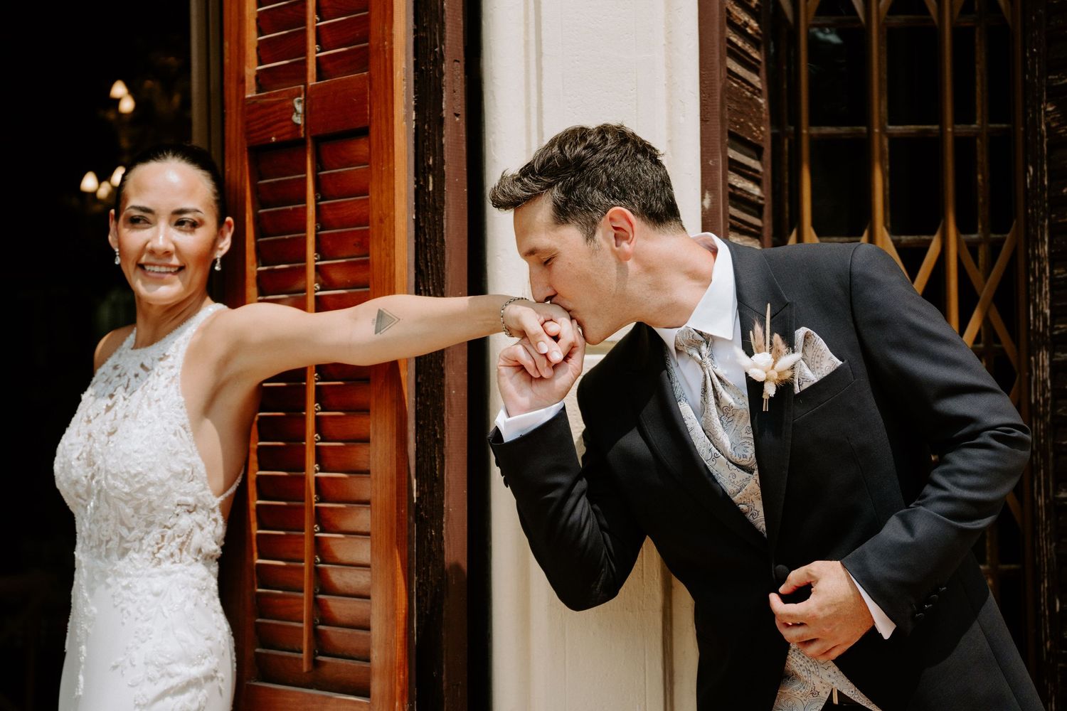 Groom in dark suit gallantly kisses bride's hand outside vintage wooden shuttered doors.