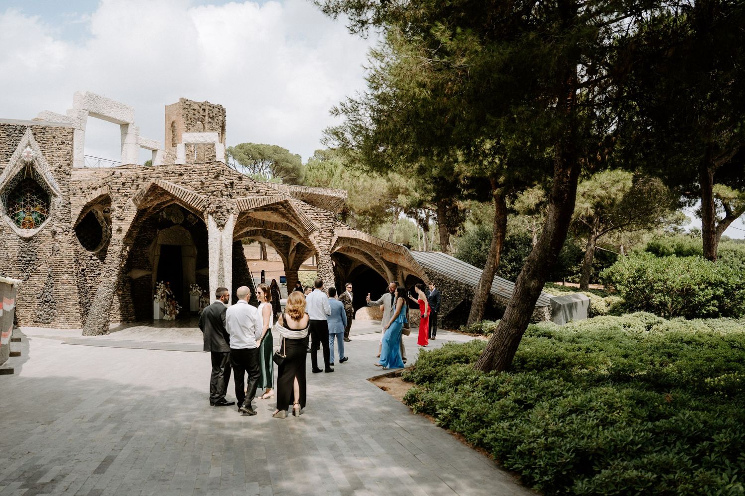 Wedding guests mingling outside a unique Gaudi-style stone architecture venue surrounded by trees.