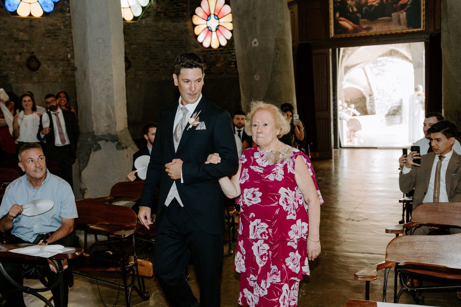 Groom in black suit escorting an older woman in floral dress down the church aisle at a wedding.