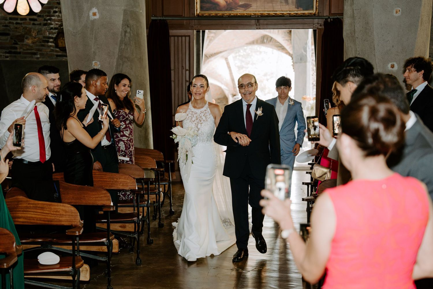 Bride in white lace gown walks down the church aisle escorted by her father among smiling guests.