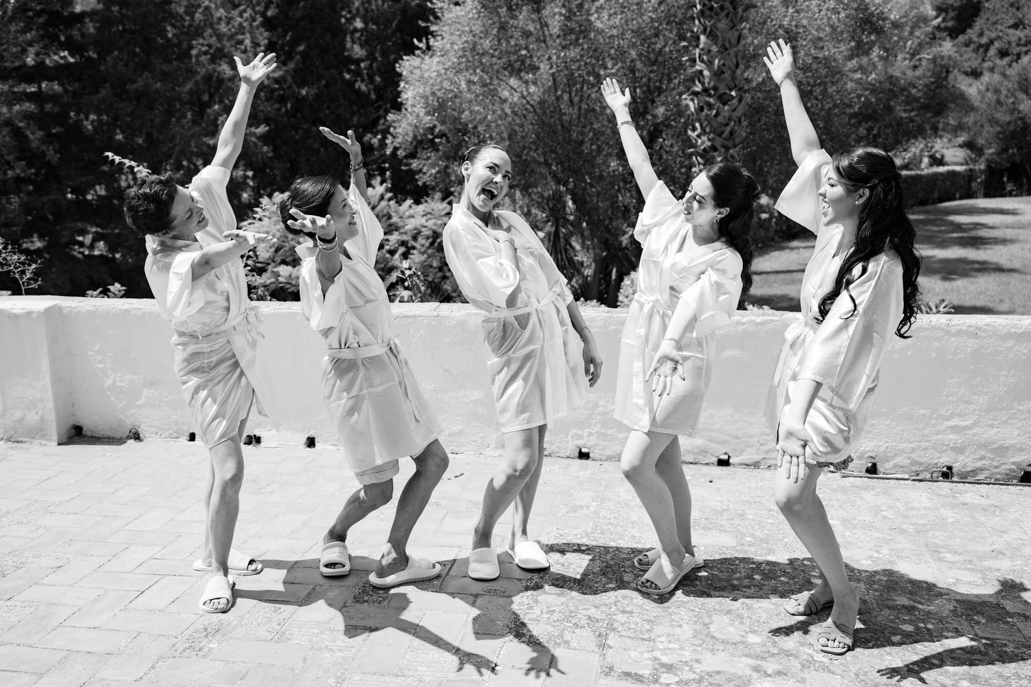 Five bridesmaids in white robes celebrate joyfully outdoors, raising arms and dancing in the sunlight.