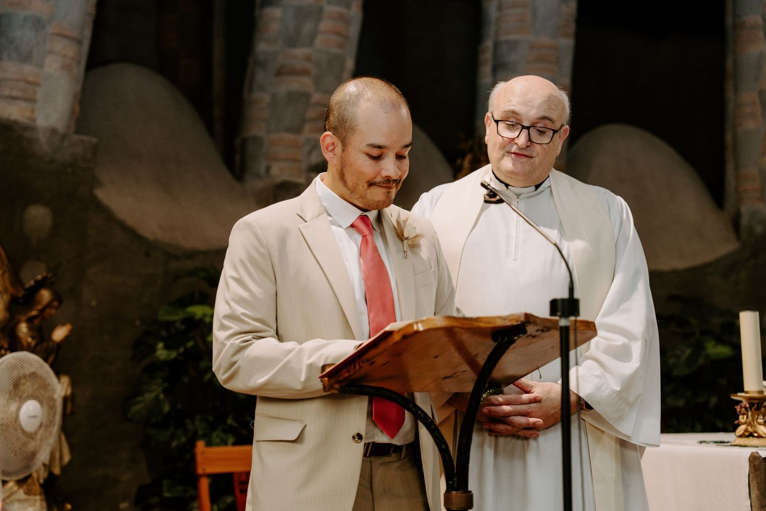 Groomsman in beige suit reads a wedding reading at the altar podium beside a priest in white robes.
