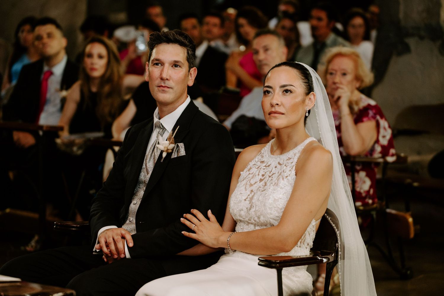 Bride and groom sit together during wedding ceremony, bride wearing lace gown and veil.
