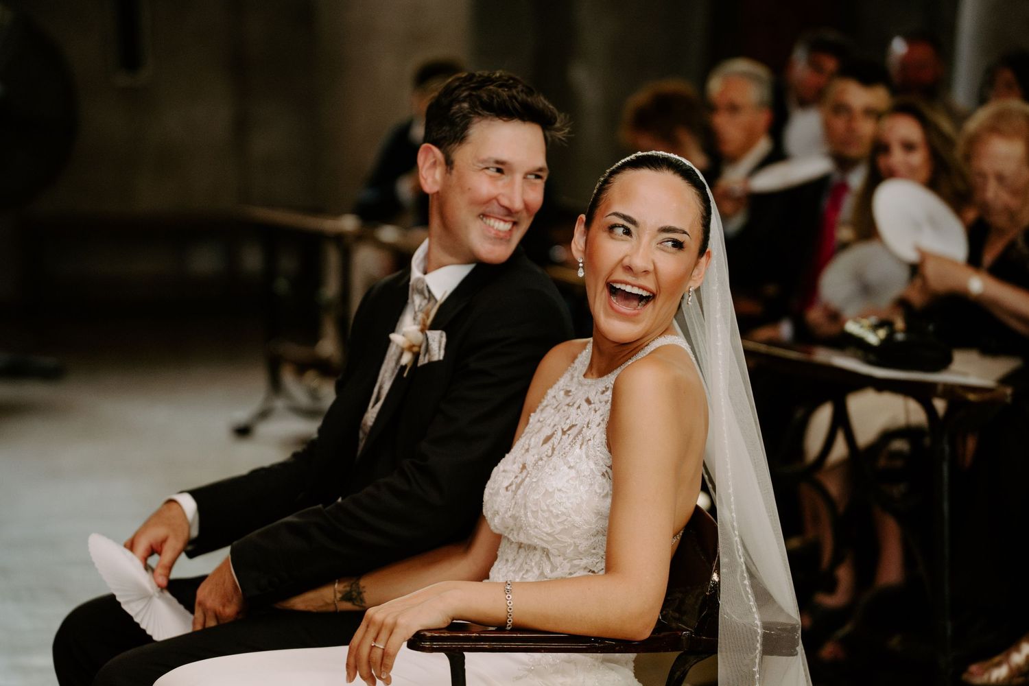 Joyful bride and groom laughing together while seated during wedding ceremony in historic venue.