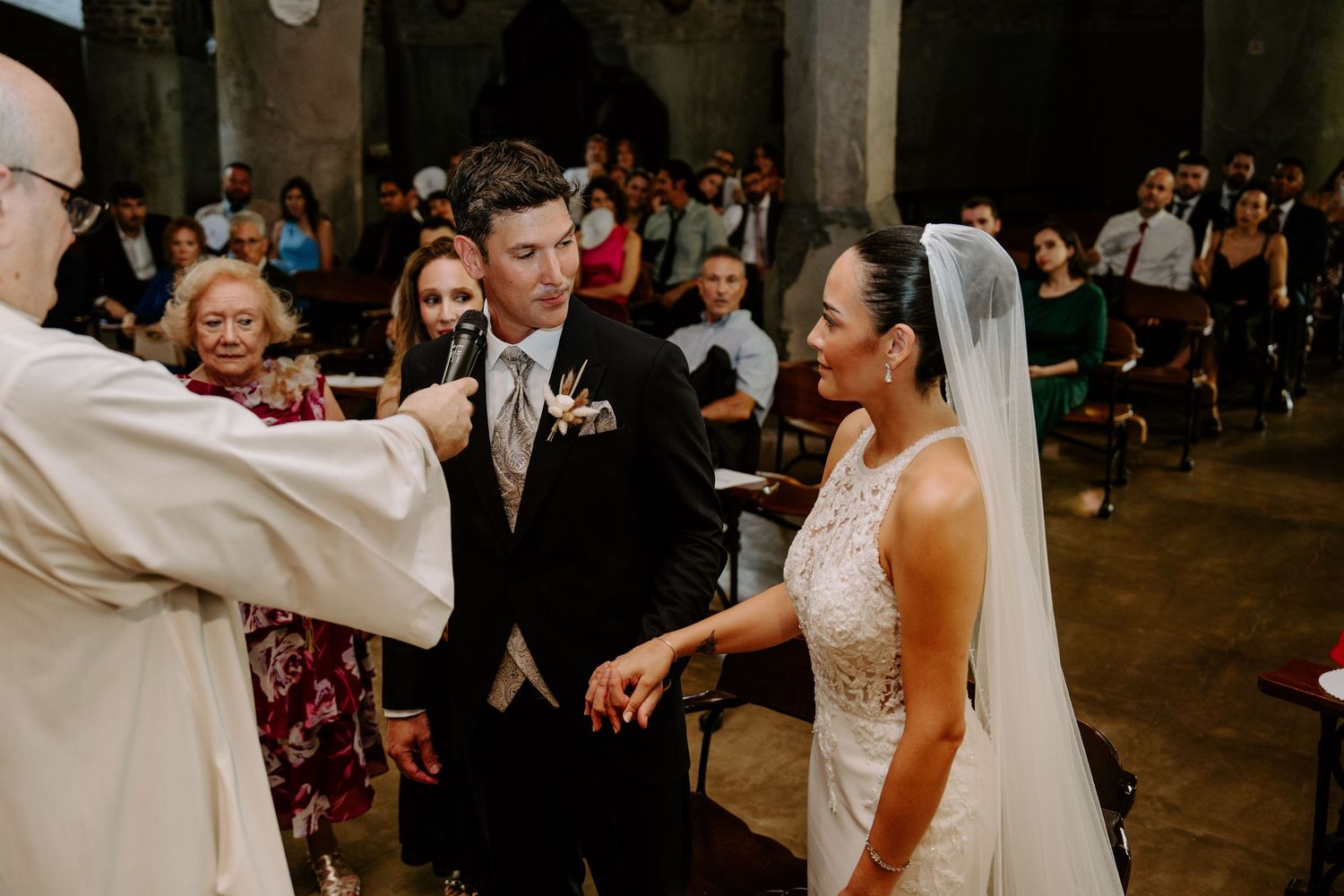 Priest officiates wedding ceremony as groom holds bride's hand at the altar in stone church.