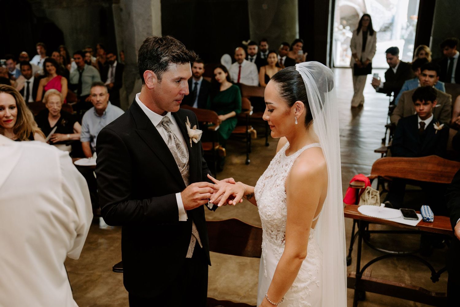 Groom places wedding ring on bride's finger during an intimate church ceremony with guests seated.