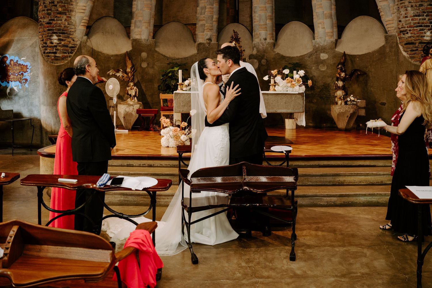 Bride and groom sharing first kiss at altar in a rustic stone chapel with wooden pews.