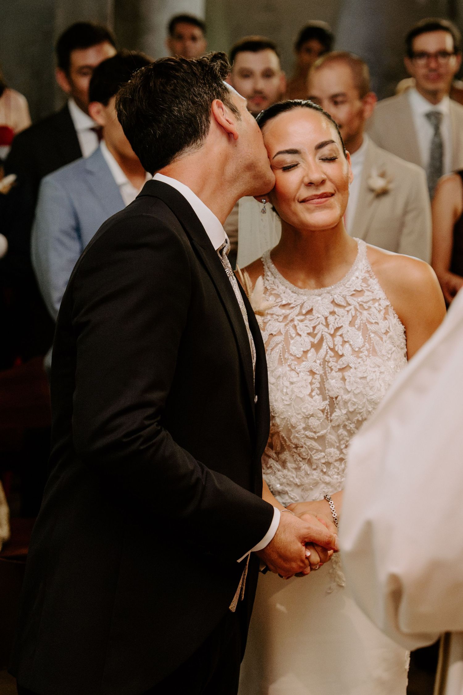Groom in black suit kisses bride on cheek during wedding ceremony as she smiles with eyes closed.