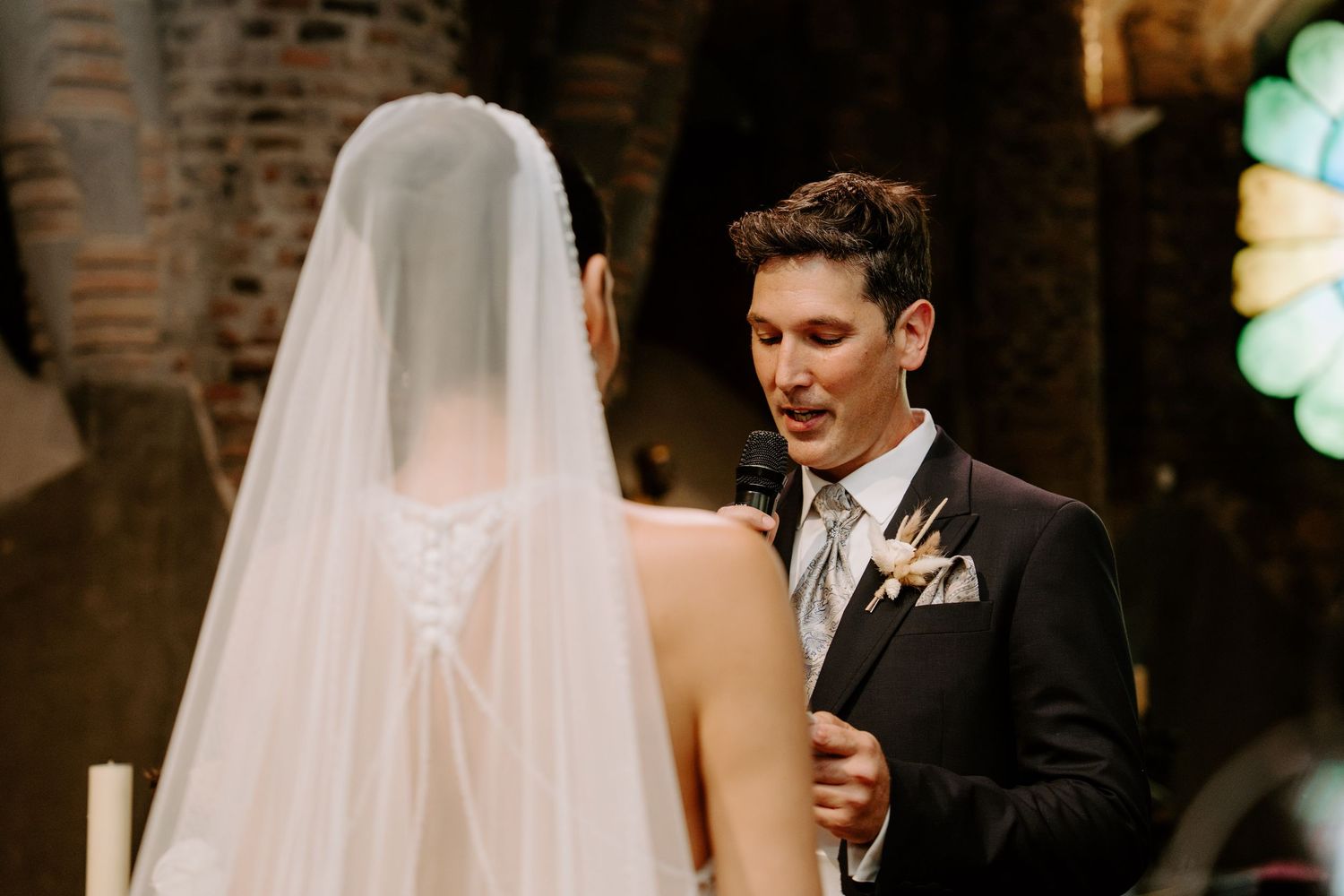 Groom gazing lovingly at veiled bride during intimate wedding ceremony in a stone church.