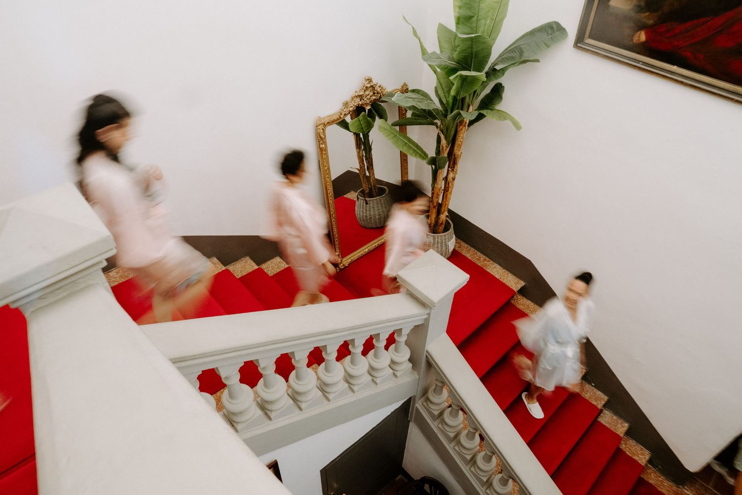 Blurred motion of bridesmaids in white dresses descending an elegant staircase with red carpet runners.