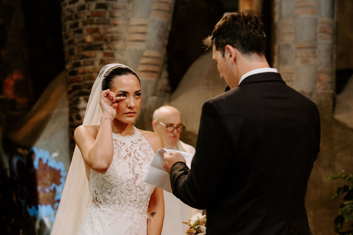 Emotional bride wipes tears during wedding vows exchange with groom in historic stone church.