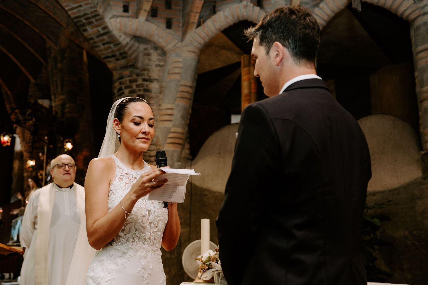 Bride in lace halter gown reads wedding vows to groom inside rustic stone church ceremony.