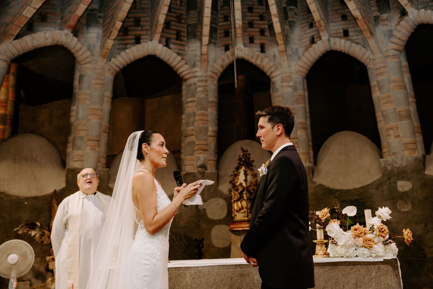 Bride exchanges vows with groom at altar inside historic brick arched church with officiant behind.