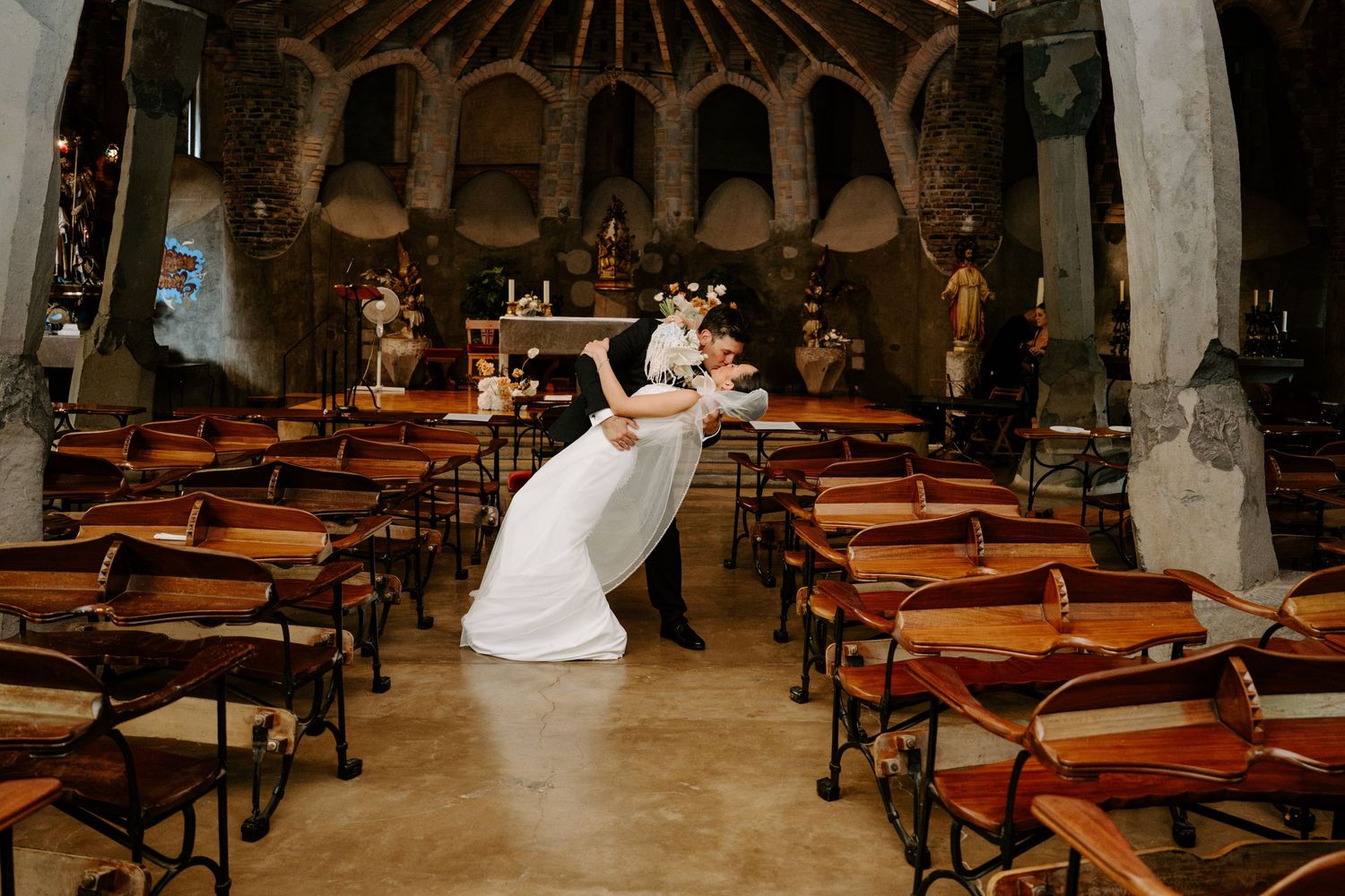 Groom dipping bride in dramatic pose inside rustic stone chapel with wooden chairs after ceremony.