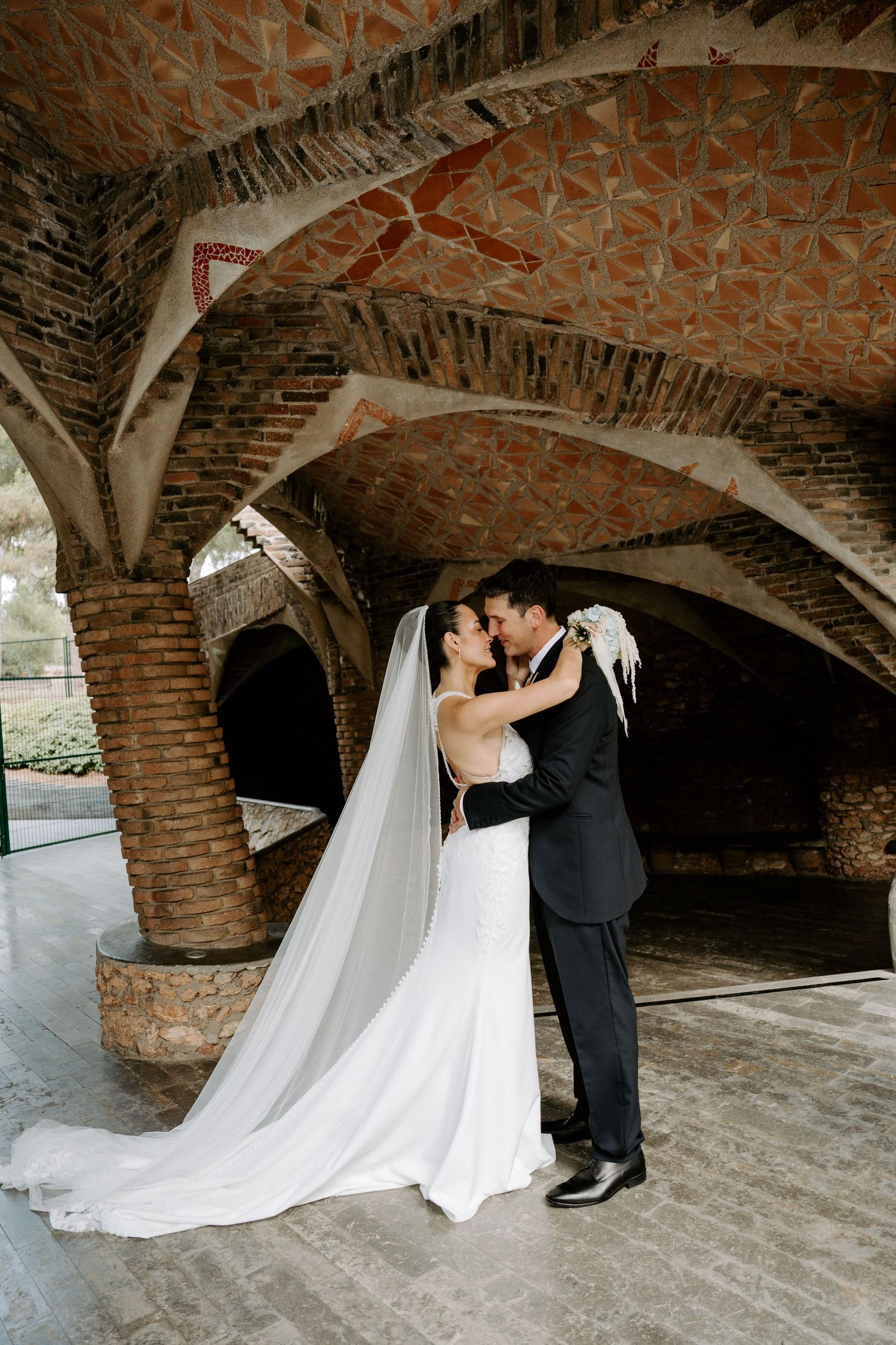 Newlyweds share romantic kiss under ornate brick arched ceiling at historic wedding venue.