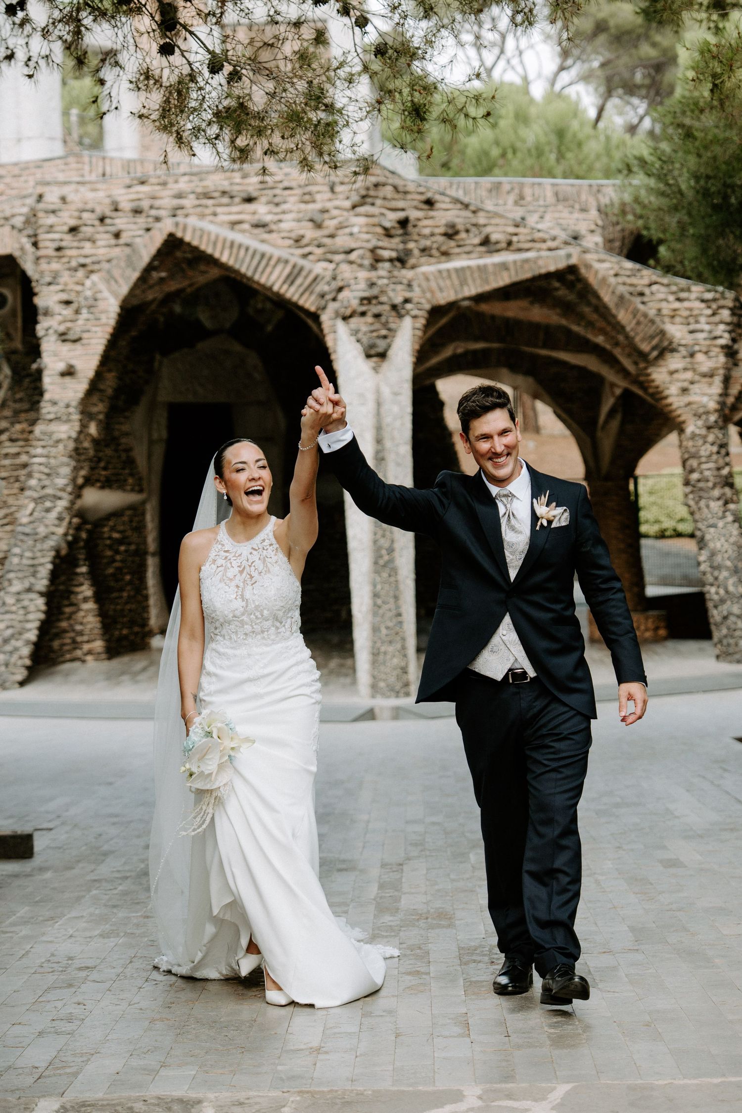 Joyful newlyweds celebrate with raised hands in front of stunning stone arched architecture on their wedding day.