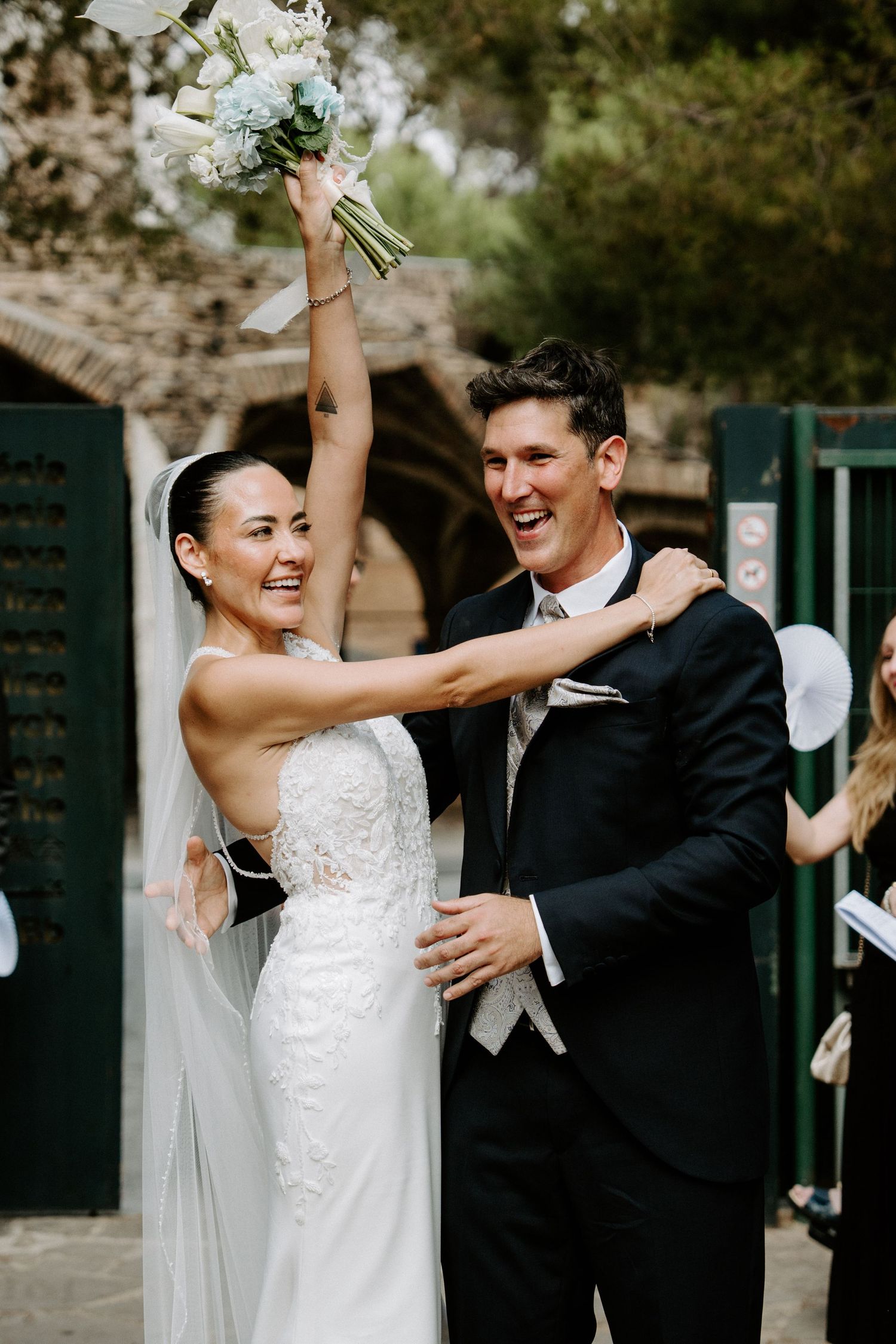 Joyful bride raising bouquet of white flowers while groom laughs beside her after outdoor ceremony.
