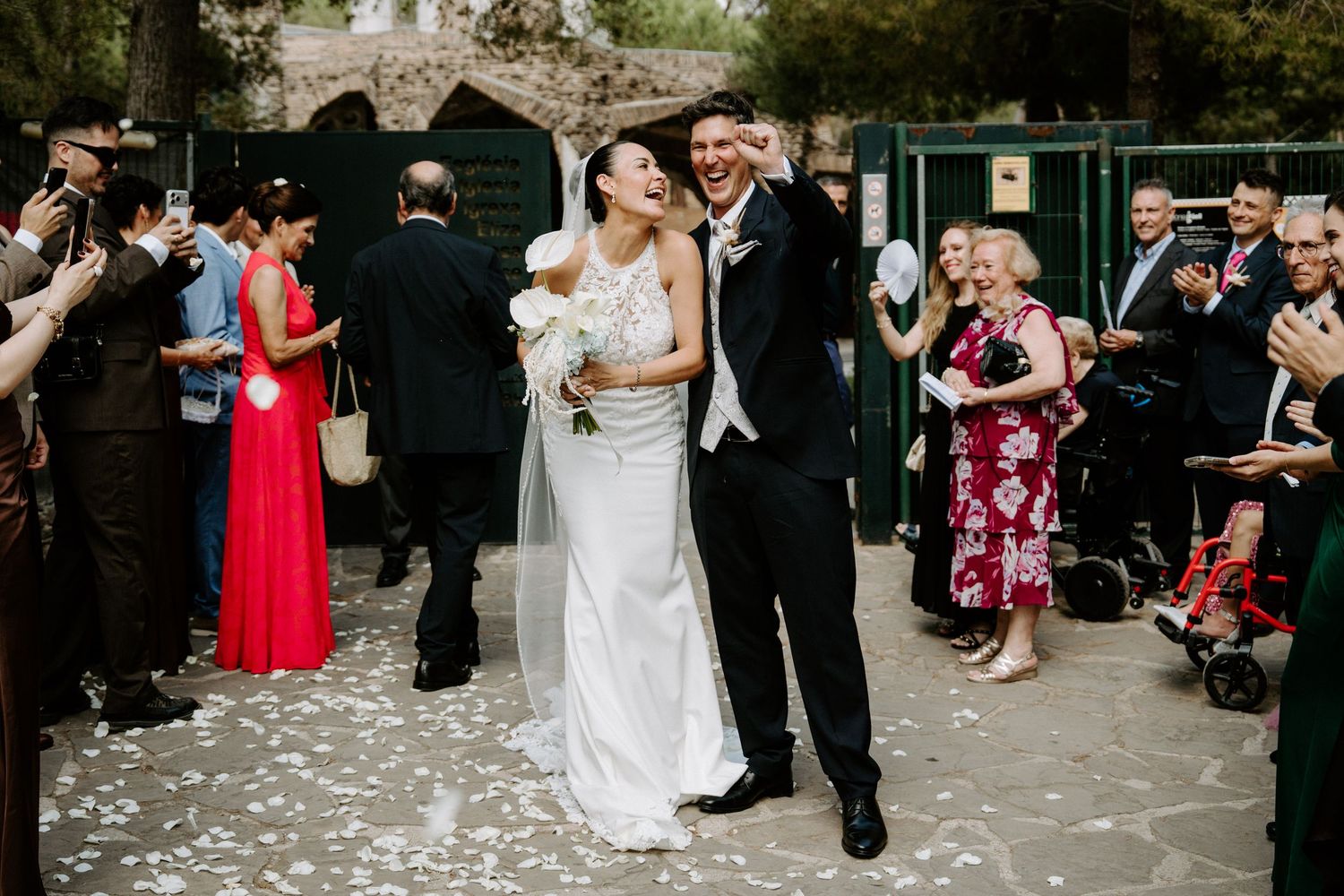 Bride and groom share a joyful kiss while guests toss white petals during their outdoor wedding ceremony exit.