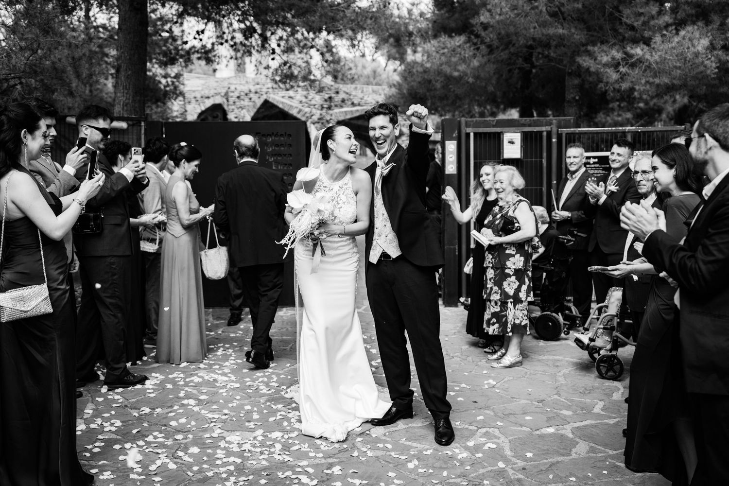 Black and white photo of newlyweds walking through confetti shower with cheering wedding guests.