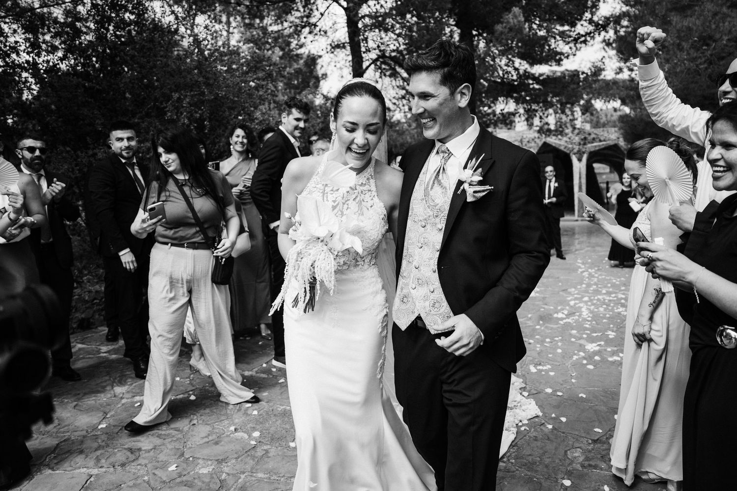 Newlyweds walk joyfully through confetti shower surrounded by cheering guests in black and white photo.