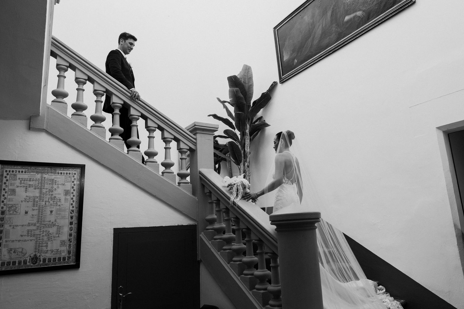 Black and white photo of bride and groom descending an elegant staircase with ornate balusters at a wedding venue.