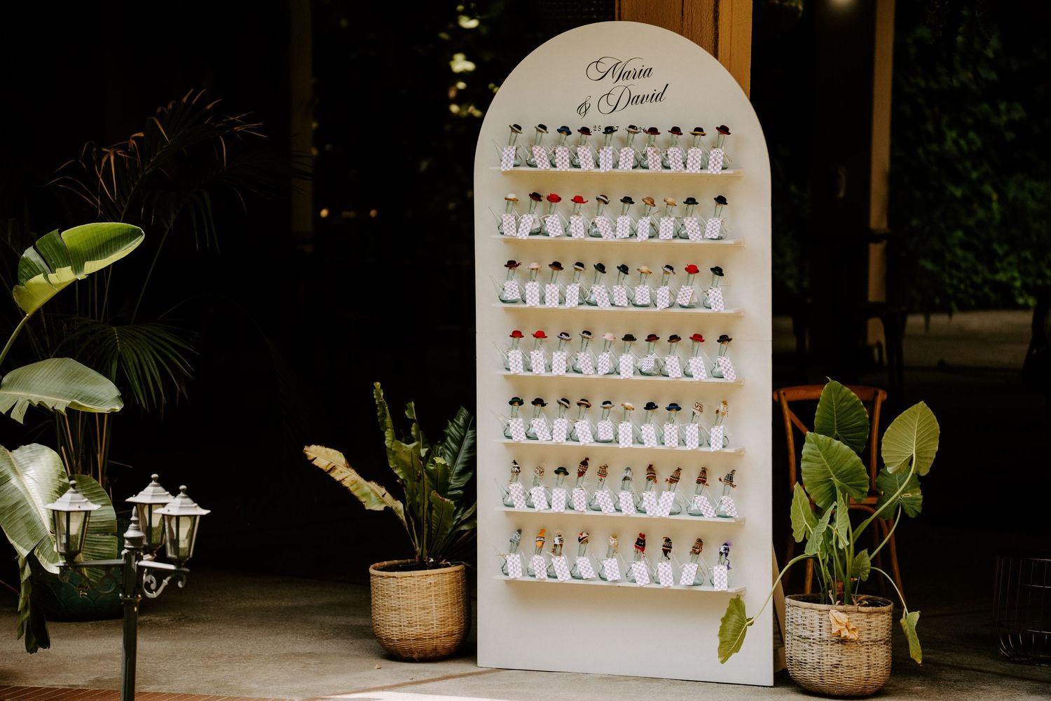 Elegant white arched wedding seating chart display with rows of escort cards and greenery in wicker baskets.