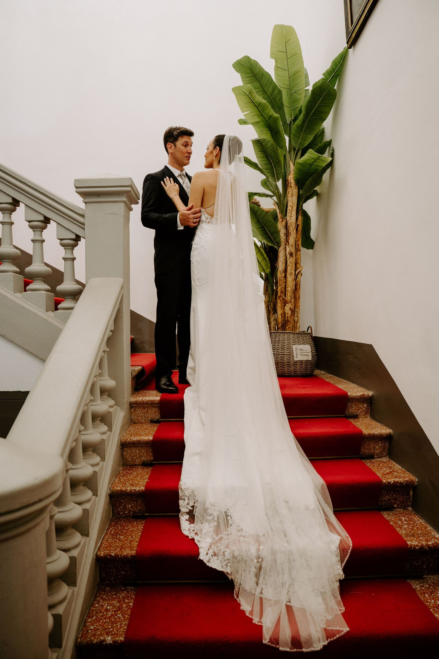 Bride and groom share a romantic moment on a red-carpeted staircase, bride wearing a long lace veil.