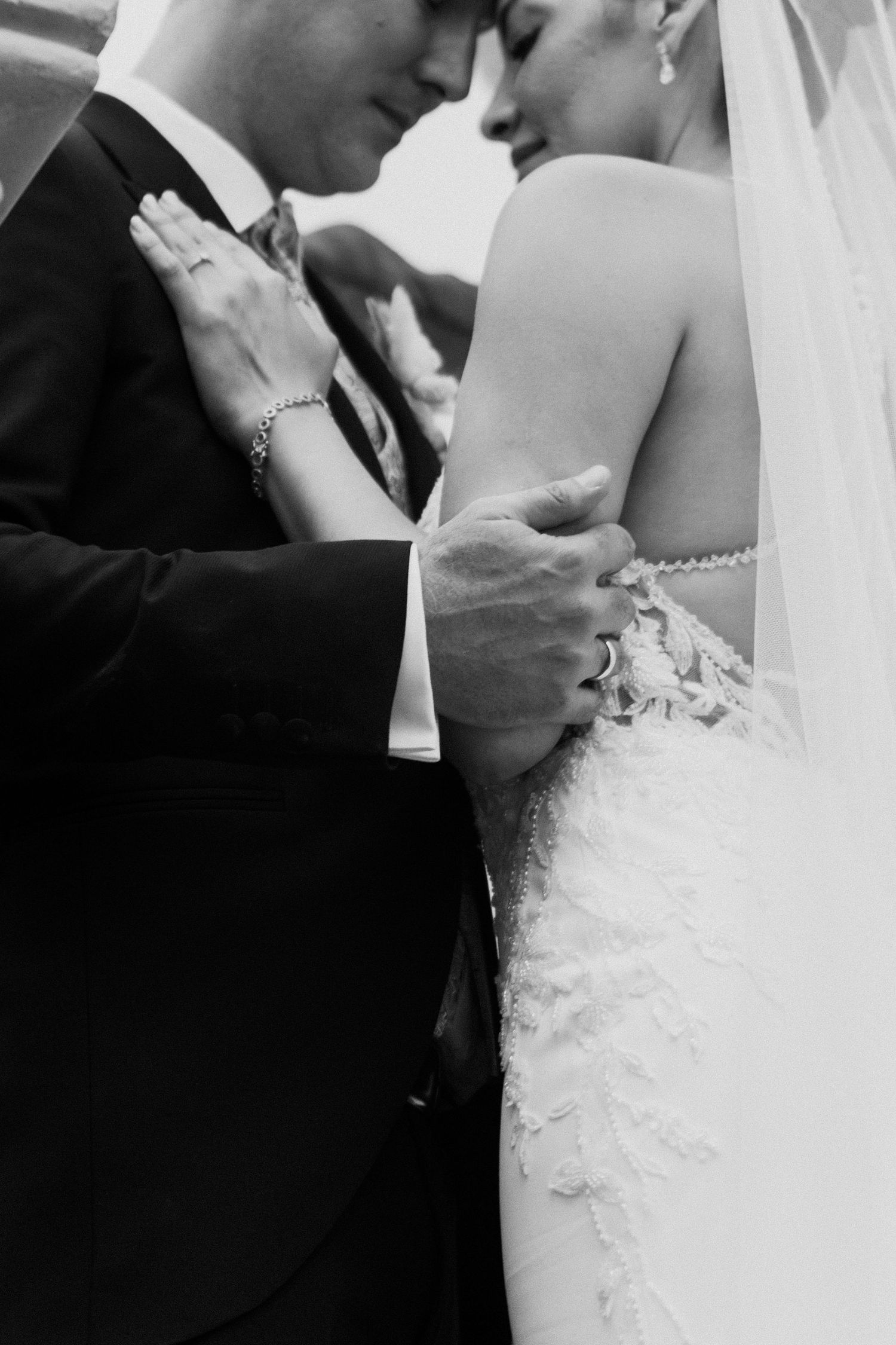 Black and white close-up of bride and groom embracing, showing lace wedding dress detail and rings.