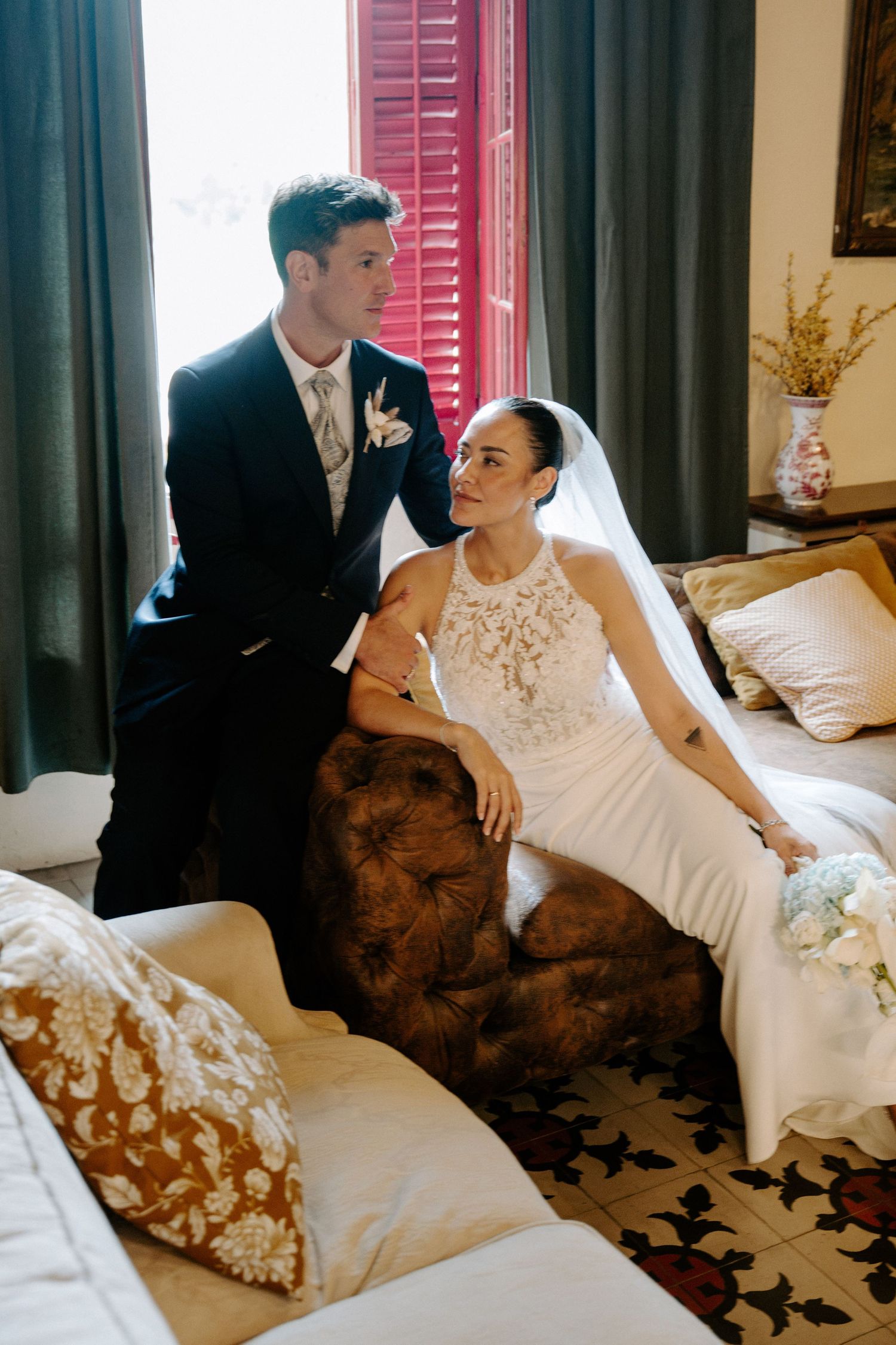Elegant bride in lace halter gown sits on vintage sofa as groom stands beside her.