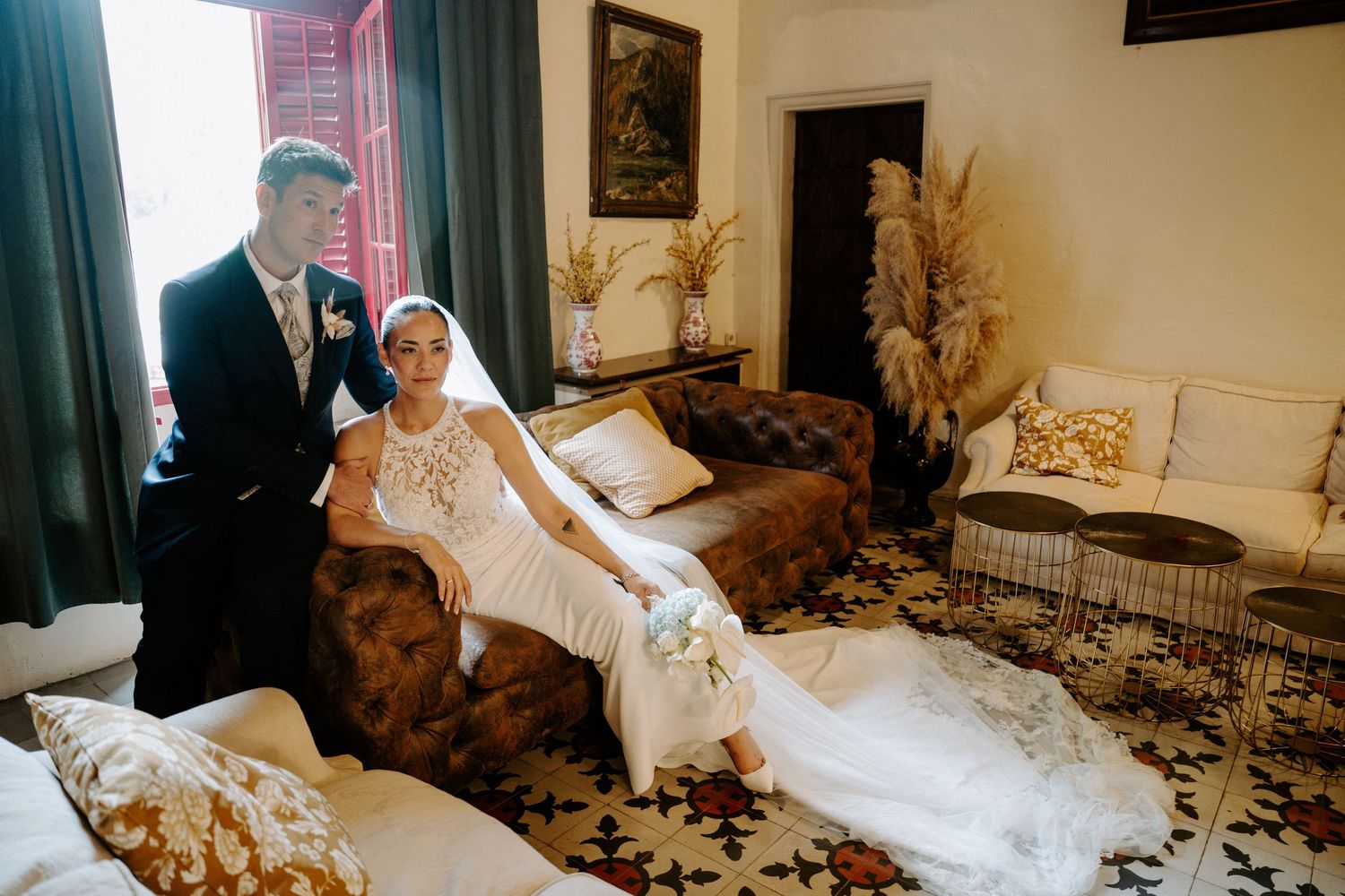 Bride in fitted lace wedding dress poses with groom on ornate sofa in vintage room.