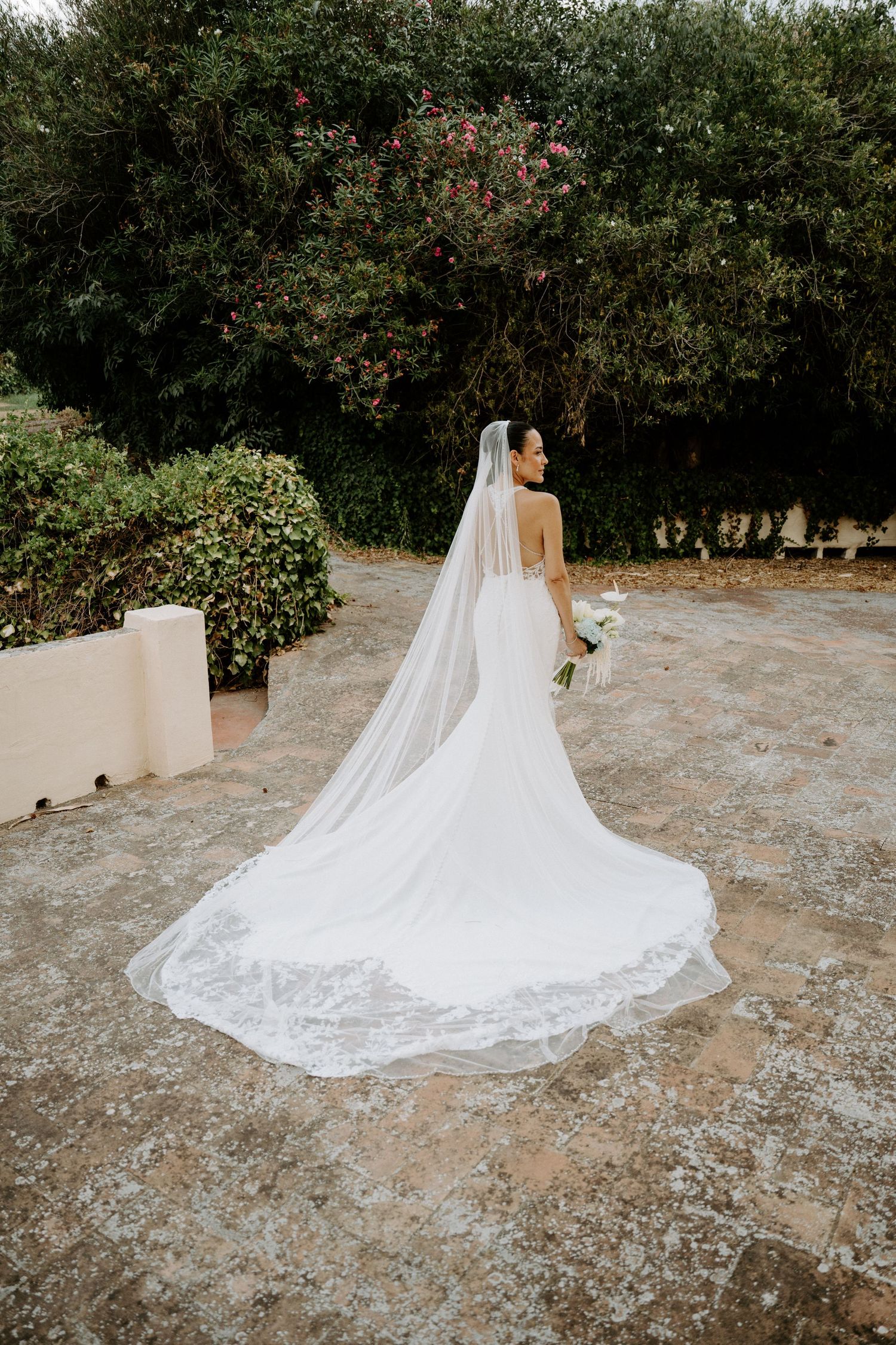 Bride in elegant backless wedding gown with long lace-trimmed cathedral veil in garden setting.