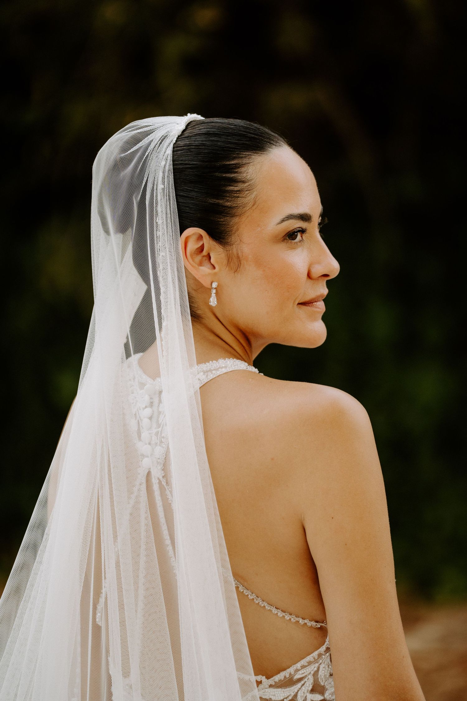 Elegant bride in profile wearing cathedral veil and halter lace gown with pearl earrings outdoors.